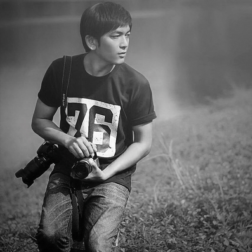 A person in a black t-shirt with the number '76' kneels on grass, holding two cameras, surrounded by misty scenery