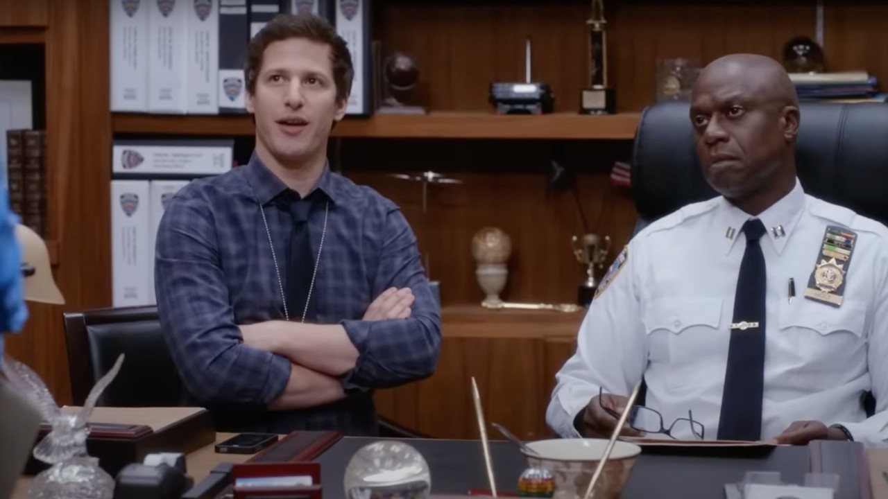 Jake Peralta and Captain Holt sitting behind Holt's desk acting suspicious while talking to Amy Santiago on Brooklyn Nine-Nine.