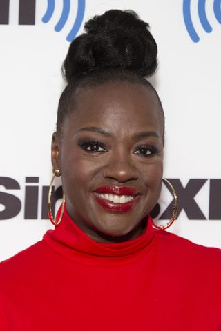 Viola Davis smiling wearing a red high-neck top, gold hoop earrings, red lipstick and her hair in a sleek high bun