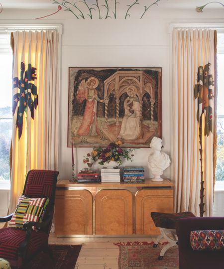 Living room with mural on ceiling, a wooden console with an antique tapestry hanging above, a classic bust and books stacked on the console, a red armchair with a geometric cushion, and a footstool. 