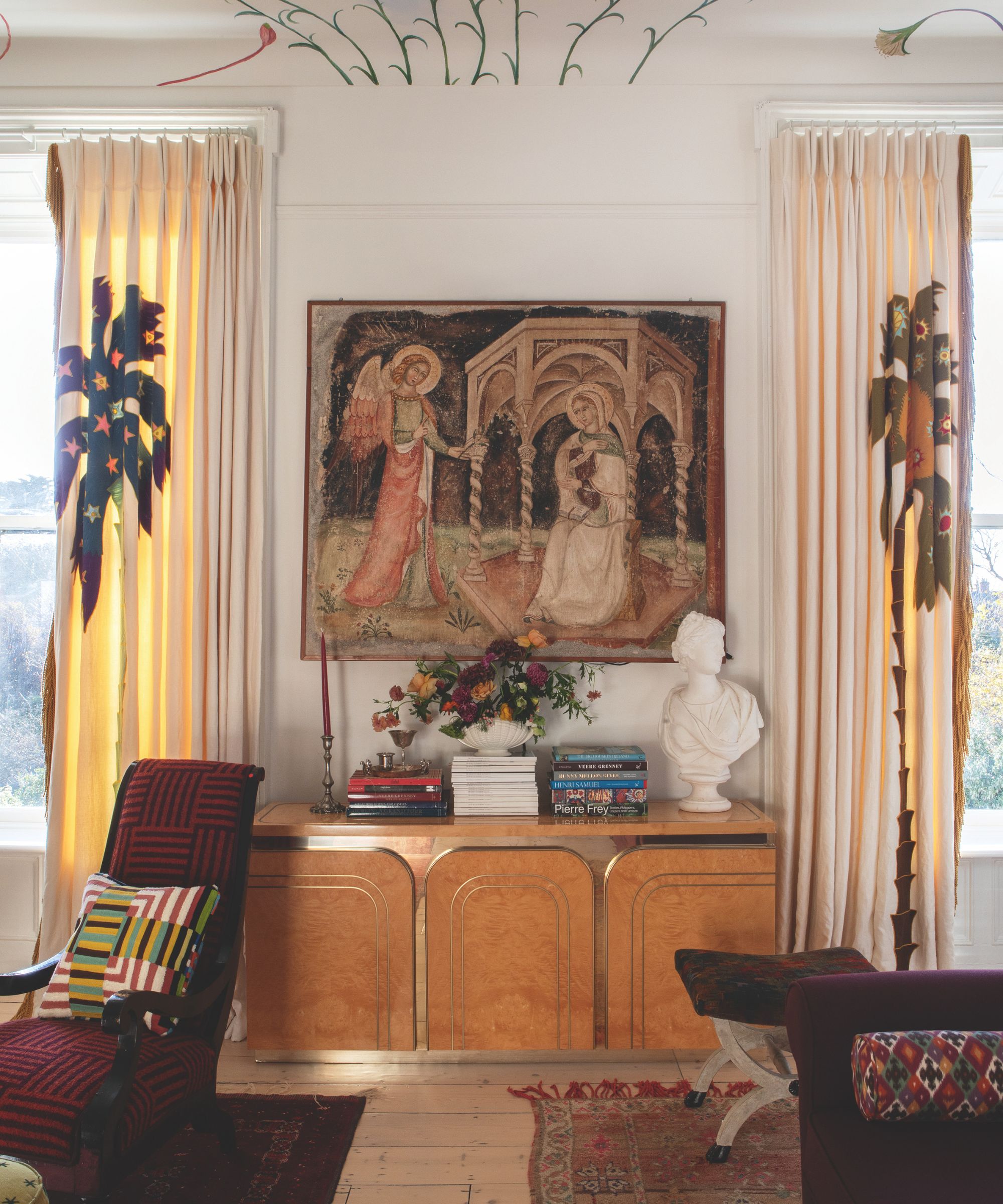 Living room with mural on ceiling, a wooden console with an antique tapestry hanging above, a classic bust and books stacked on the console, a red armchair with a geometric cushion, and a footstool.