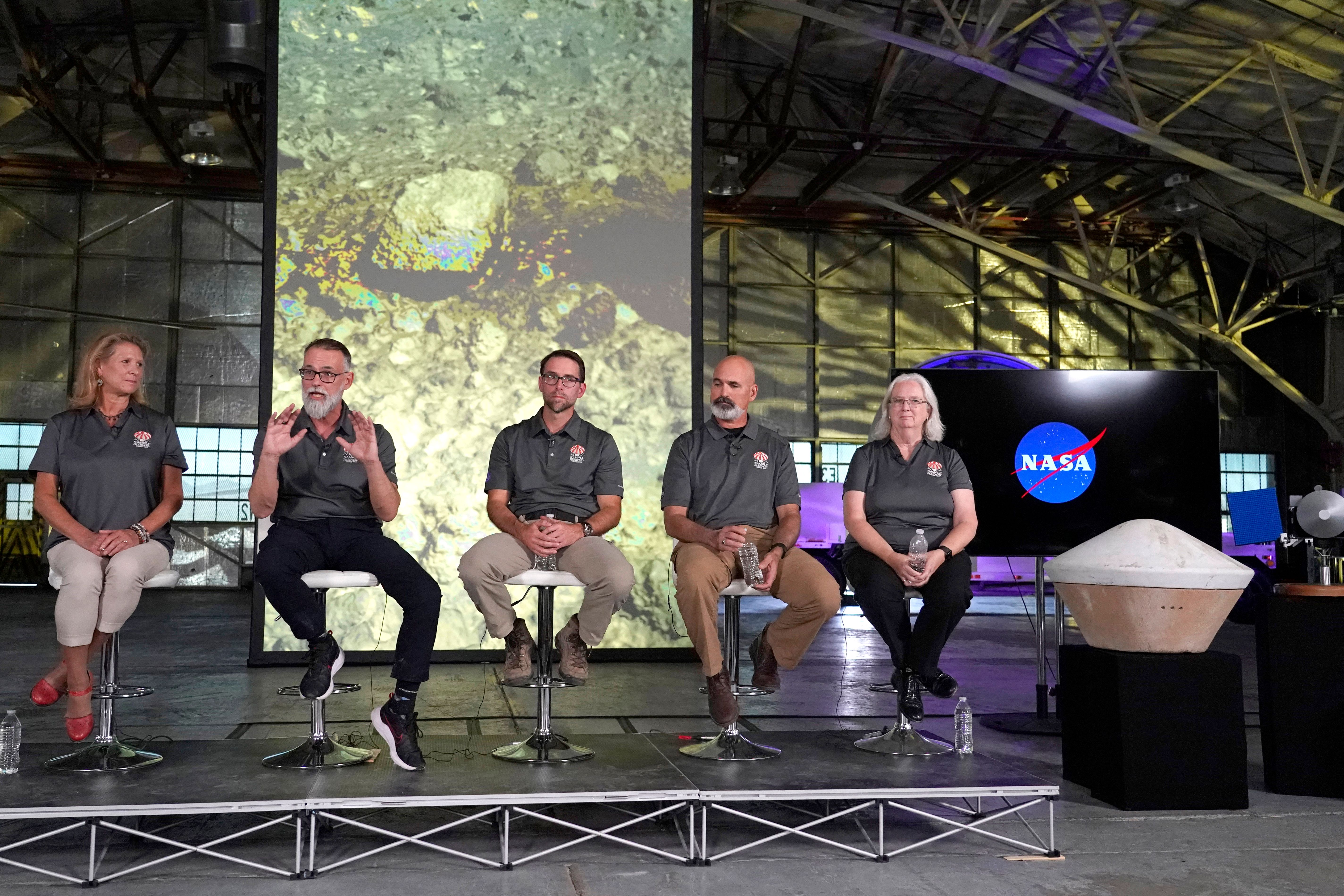 five people in matching grey shirts sit on a podium in an aircraft hangar speaking to a crowd