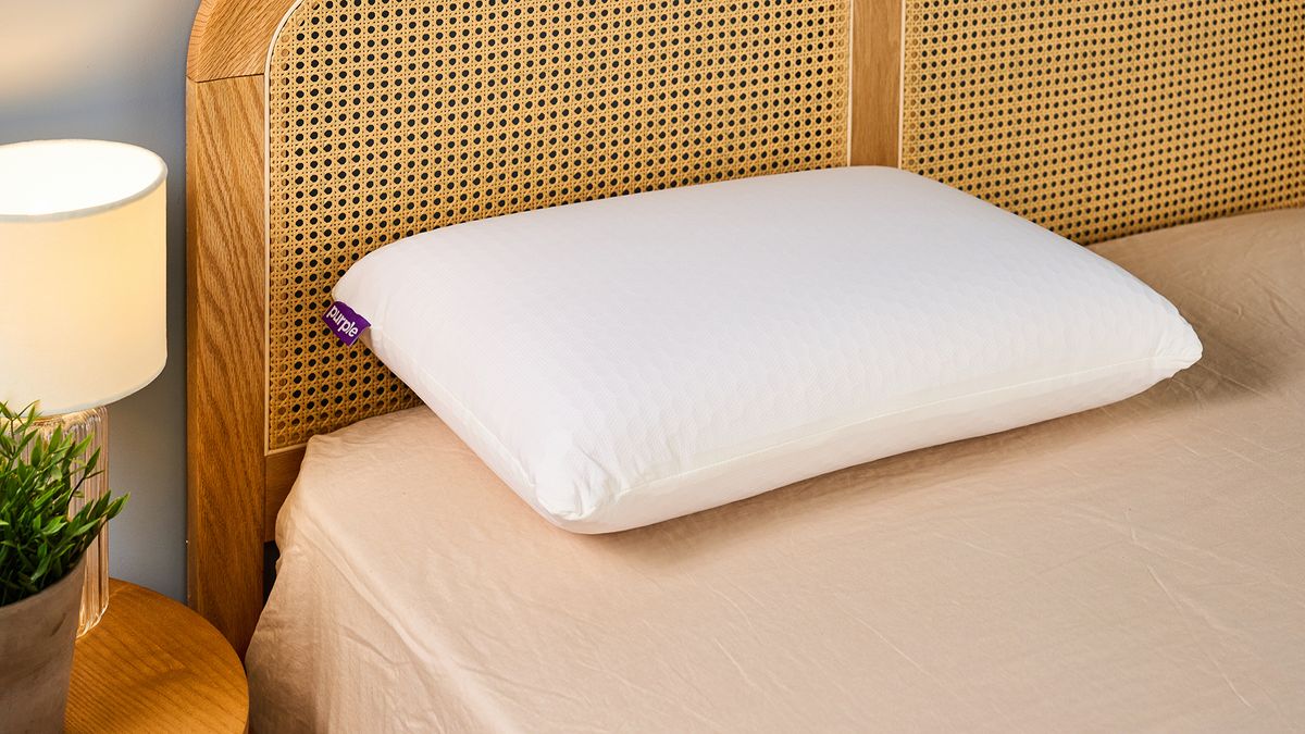 A white pillow placed on bed against a rattan headboard next to a side table with a lamp and potted plant on top
