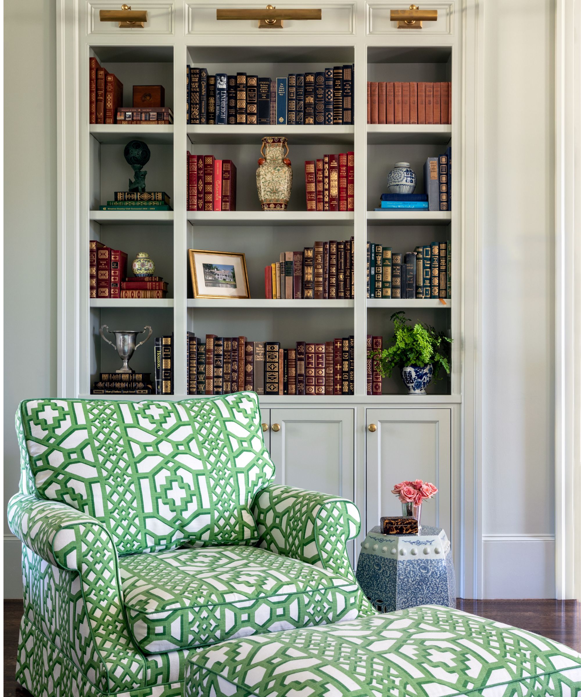 white bookshelves with cupboards below with brass picture lamps styled with vintage booked with a green lattice print chair and footstool