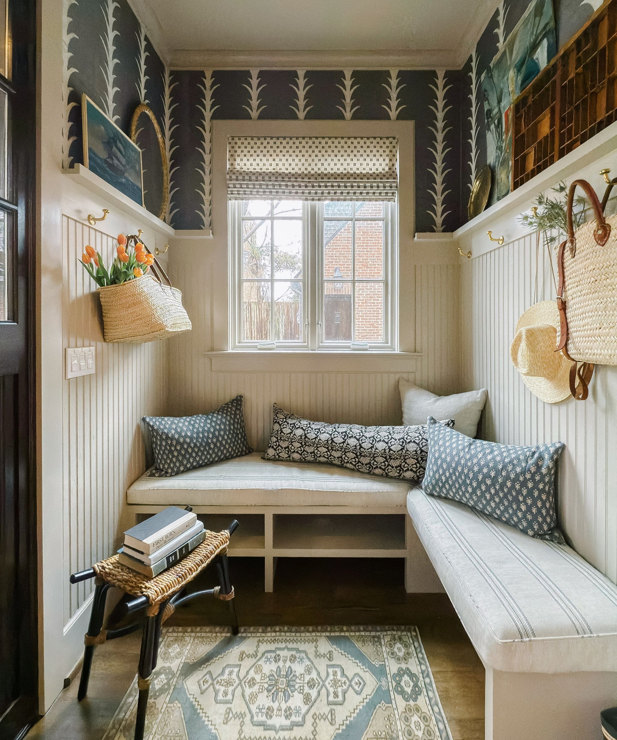 Half wallpapered reading room with built-in bench with striped cushion, small Persian rug, wall panelling with ledge and coat hooks, and an array of blue and white floral cushions