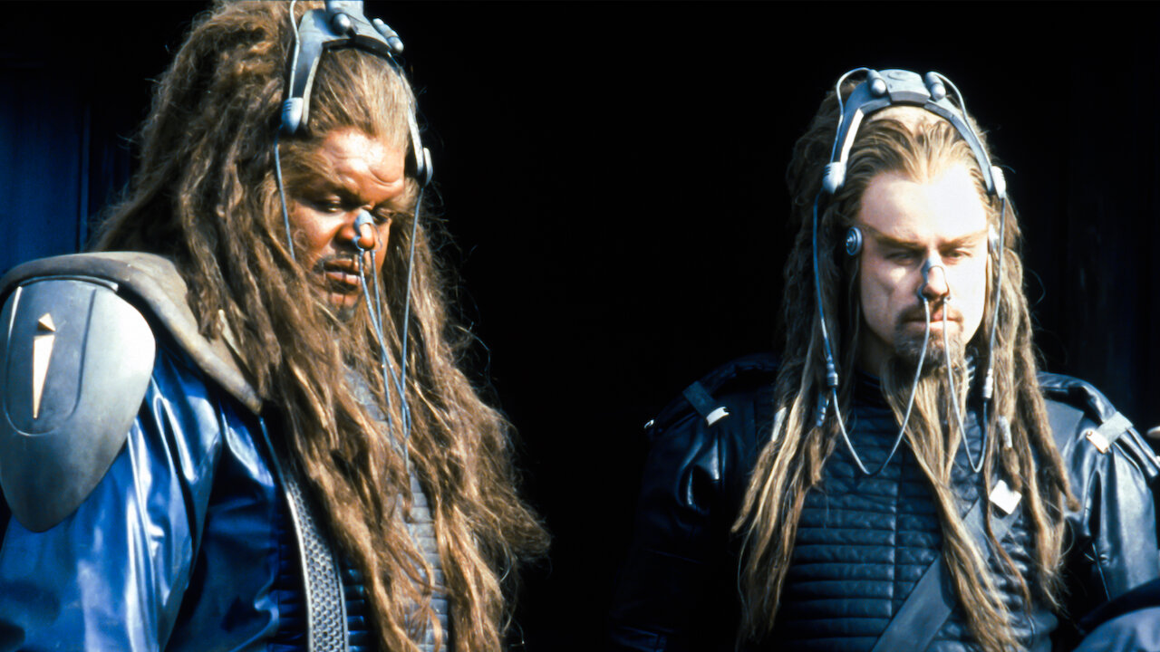 A weird alien with dreadlocks lifts a smaller human tribesman