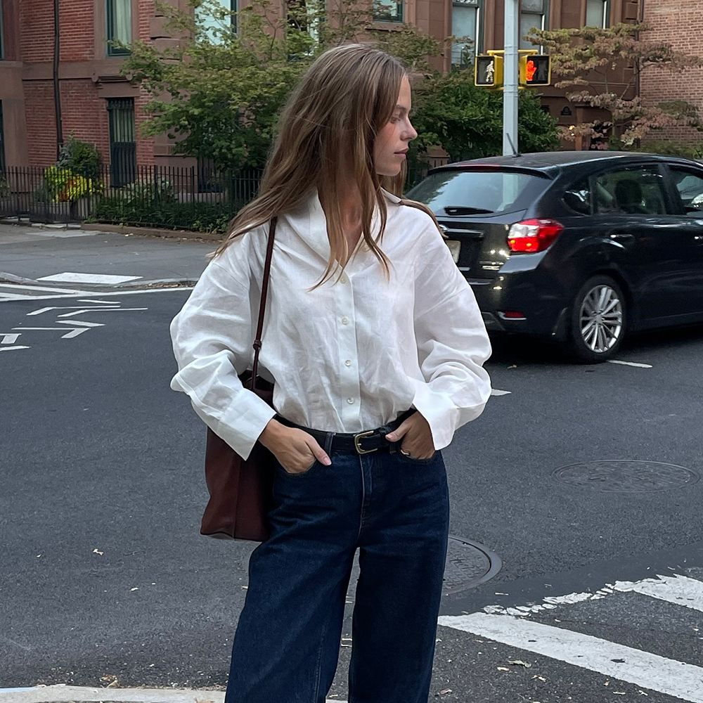 a woman wearing a white button-down, blue jeans, burgundy bag, and black flip flops