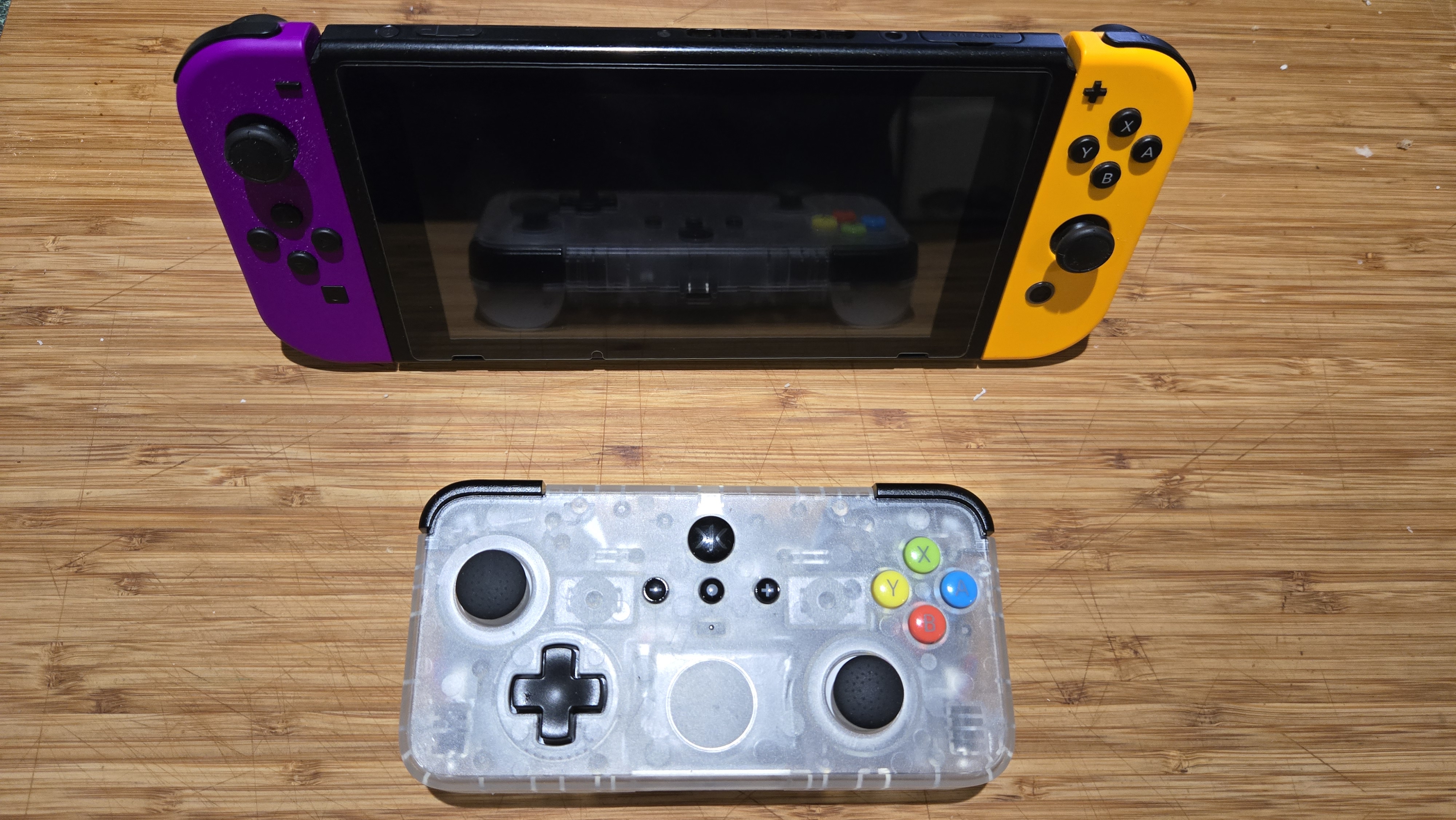 A Classic Clear edition of the CRKD NEO S rectangular controller sits upon a wooden countertop, placed beside a Nintendo Switch game console with docked Joycon controllers. The CRKD controller is half the size of the Nintendo Switch's screen, but not as small or as light weight as the Joycons. The body of the CRKD controller is made out of a cloudy clear plastic that allows the viewer to see the internals.