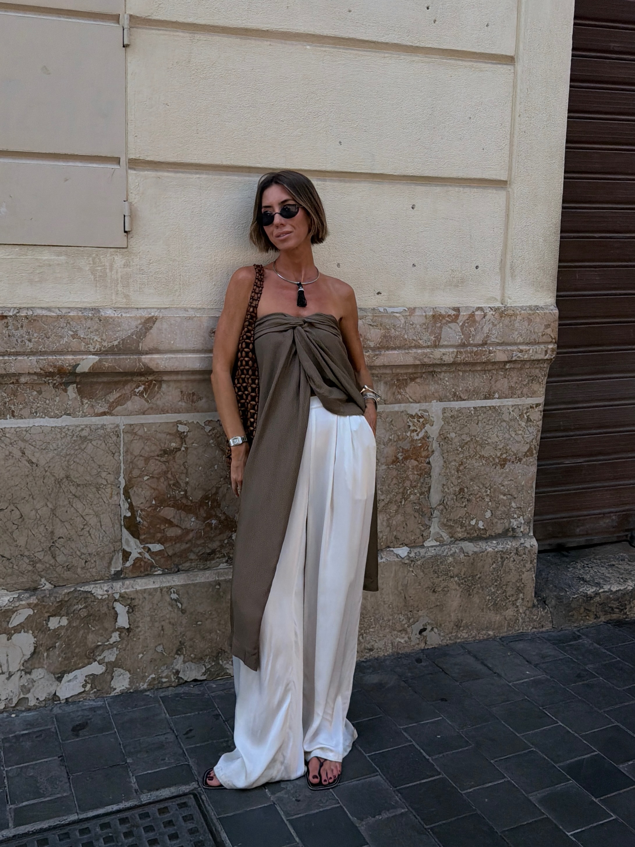 An image of an influencer in a long, strapless taupe top and white satin trousers. She wears flips flops and holds a brown beaded bag.