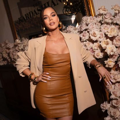 a brunette woman wearing a light brown, leather mini dress and a beige blazer in front of a floral setup
