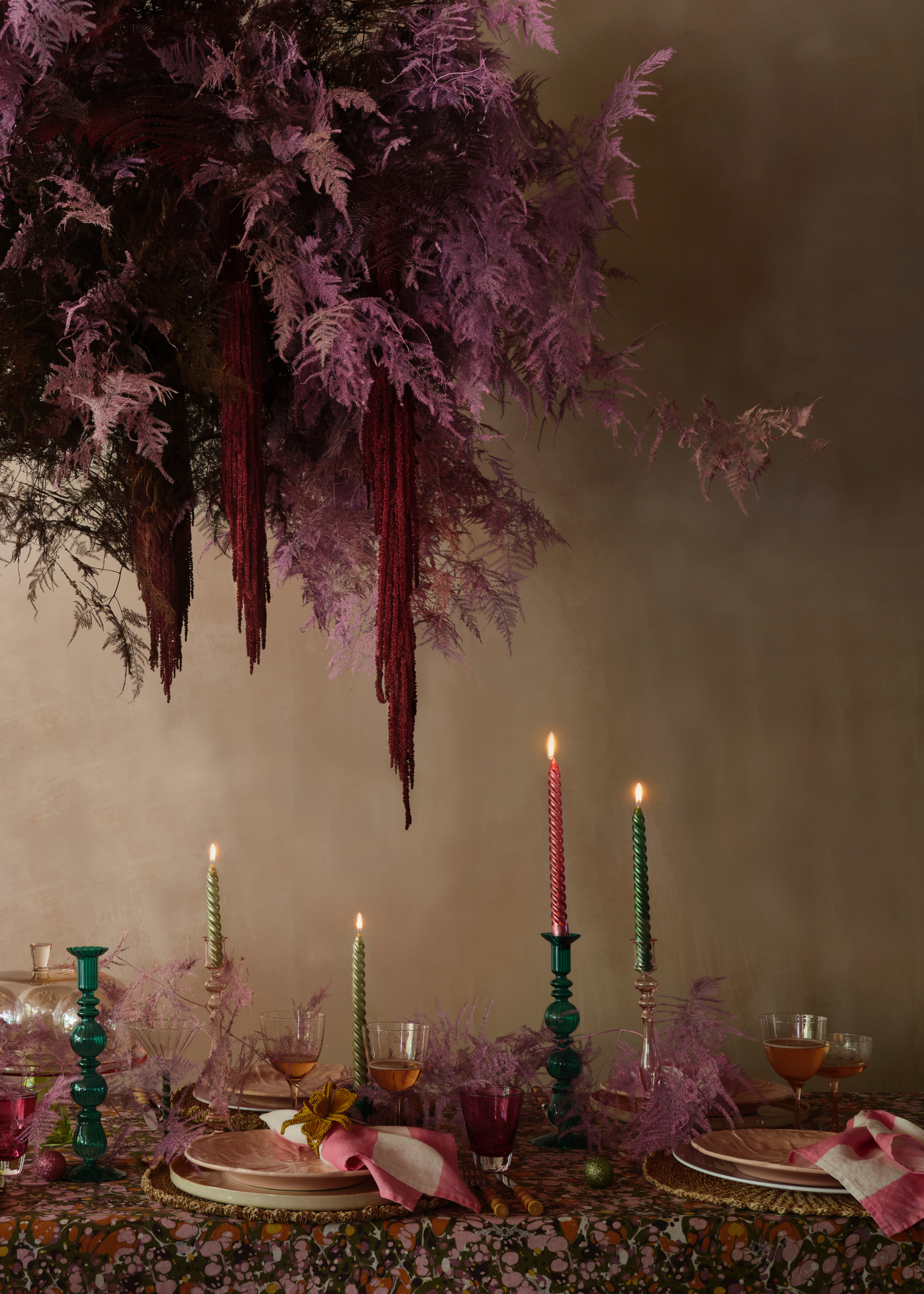A foliage display in a purple hue with pops of draping red, hanging above a table with multi-colored lit taper candles at varying heights in amongst table settings with napkins