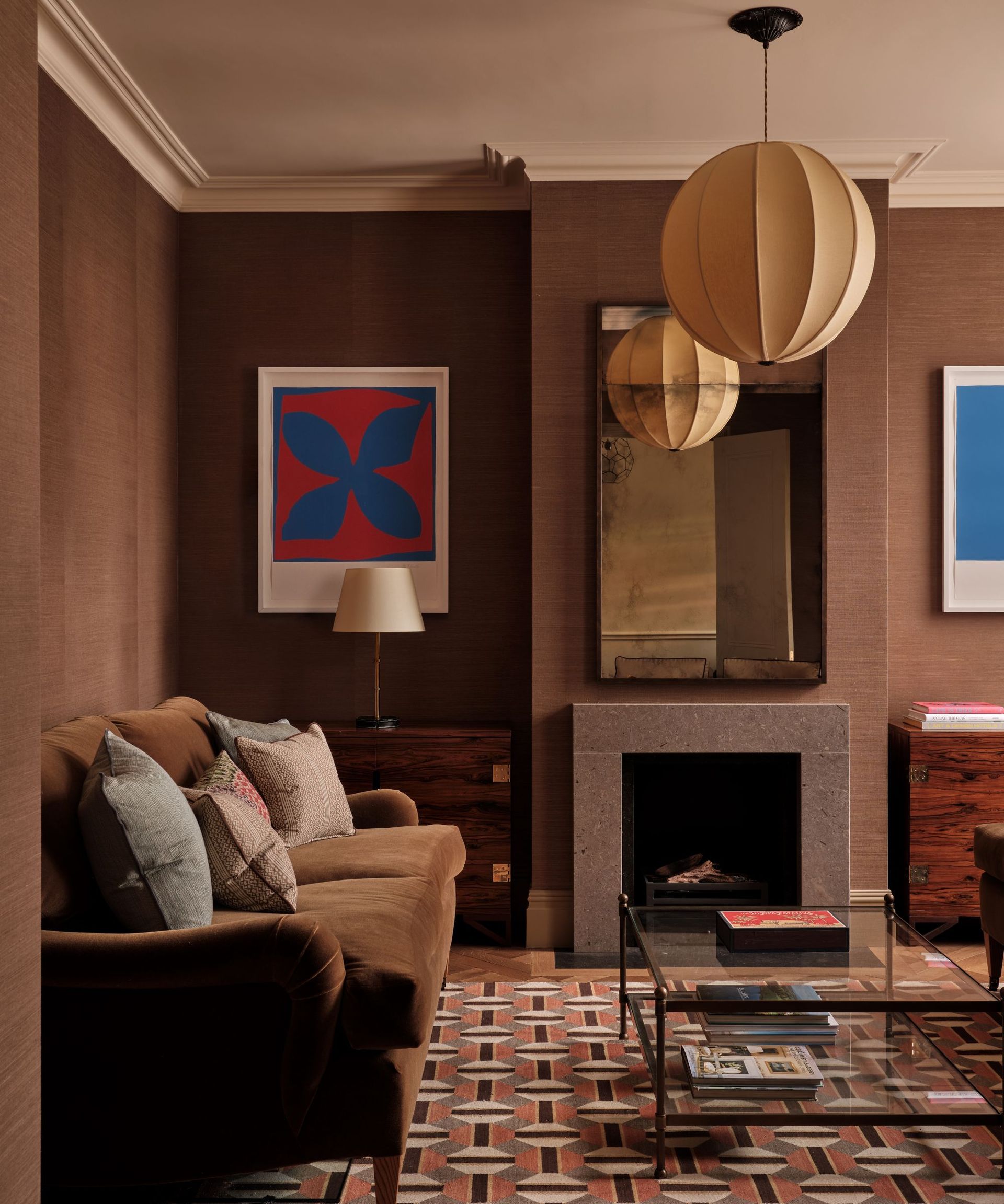 Living room with brown grasscloth walls, cream ceiling, lantern pendant light, geometric orange and brown rug, glass coffee table, brown velvet sofa, and a fire