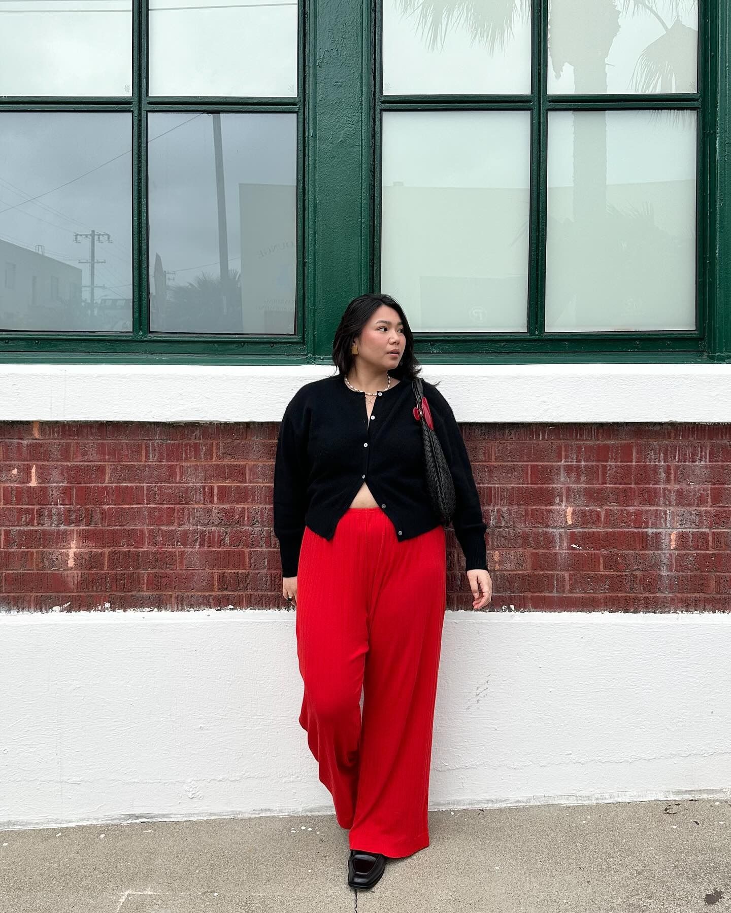 Influencer wears red trousers with square-toe boots and a black cardigan, styled partially unbuttoned.