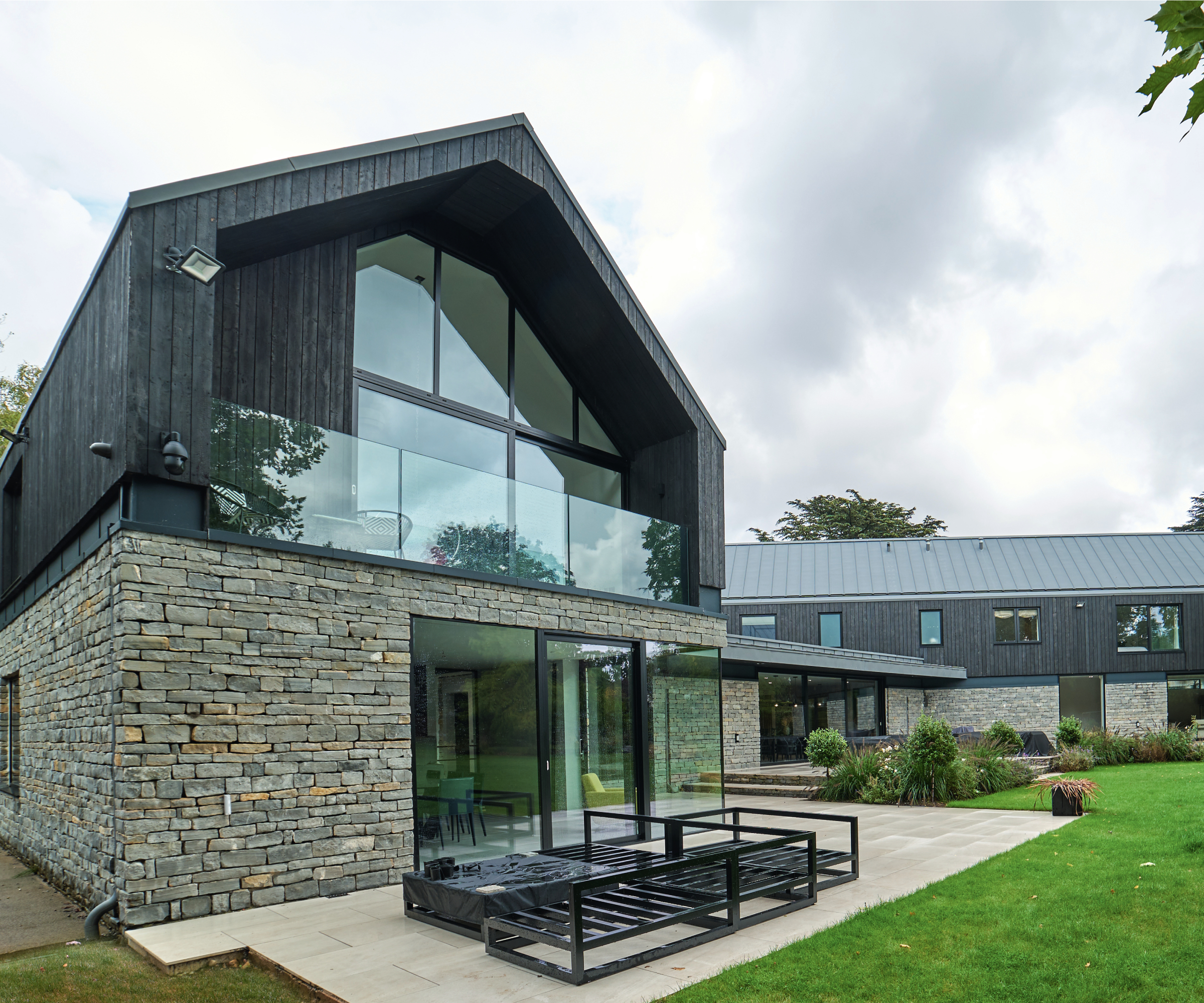 modern house with grey stone ground floor featuring patio doors with corner glazing, pitched gable end second floor with large windows and glass balustrade