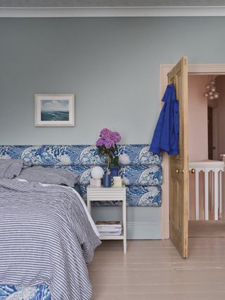 light blue bedroom with blue and white upholstered headboard, striped blue bedding, a white side table with purple flowers, a yellow door and baby pink floorboards