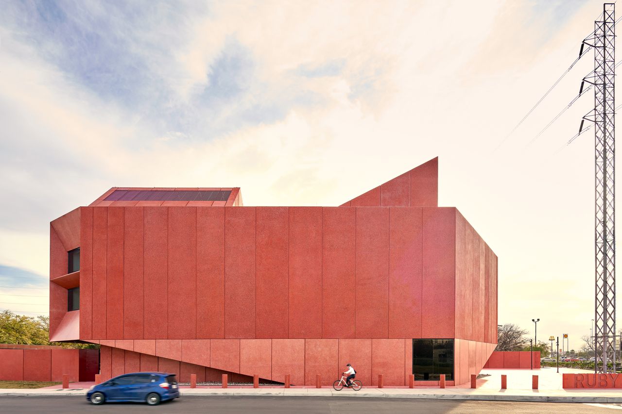 Daytime image of the new contemporary Art Centre, red exterior, road, blue car, cyclist, surrounding trees, electricity pylon, blue cloudy sky 