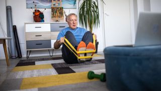 An older man works out at home. He's sat on a rug, facing a laptop screen that's open. He uses a resistance band, looped around his feet, and is pulling against it. Near him is a small green dumbbell.