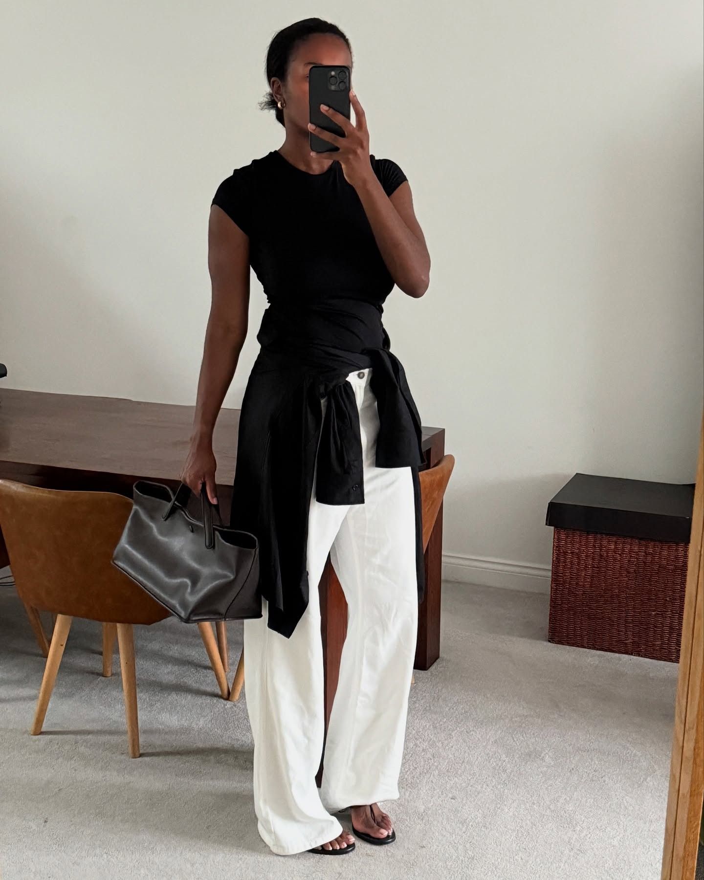 British fashion influencer Marilyn Nwawulor-Kazemaks poses for a mirror selfie in her chic London dining room wearing a black fitted T-shirt, a black button-down tied around her waist, black The Row top handle tote bag, baggy white jeans, and heeled black thong sandals