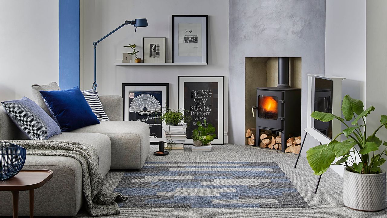 A grey living room with grey and blue carpet tiles, blue floor lamp, framed wall art on floor and floating shelves, a traditional black woodburning stove in concrete alcove and potted houseplant