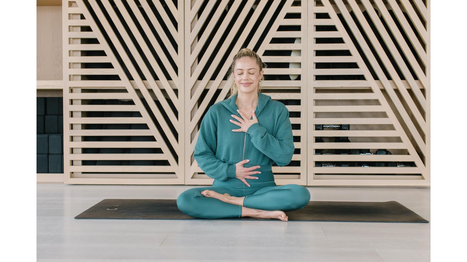woman dressed in jade green hoodie and legging sitting cross legged in a meditative position with a hand on her chest and abdomen, facing the camera eyes closed.