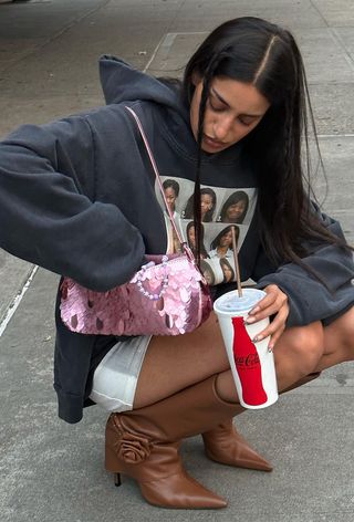 A woman crouching in the street wearing a gray graphic sweatshirt with a white miniskirt, brown knee-high boots, and a pink sequin bag.