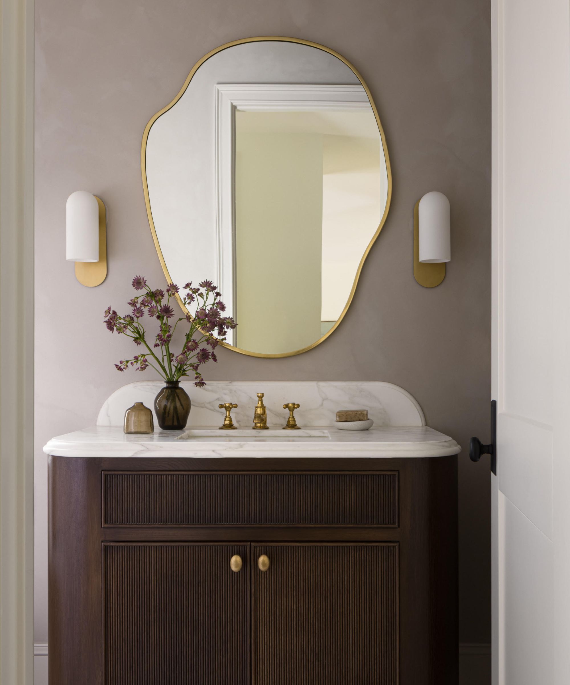 A bathroom with taupe limewash walls, a dark wooden vanity with a white sink, an organic form mirror, two modern wall sconces, and a vase of purple flowers.