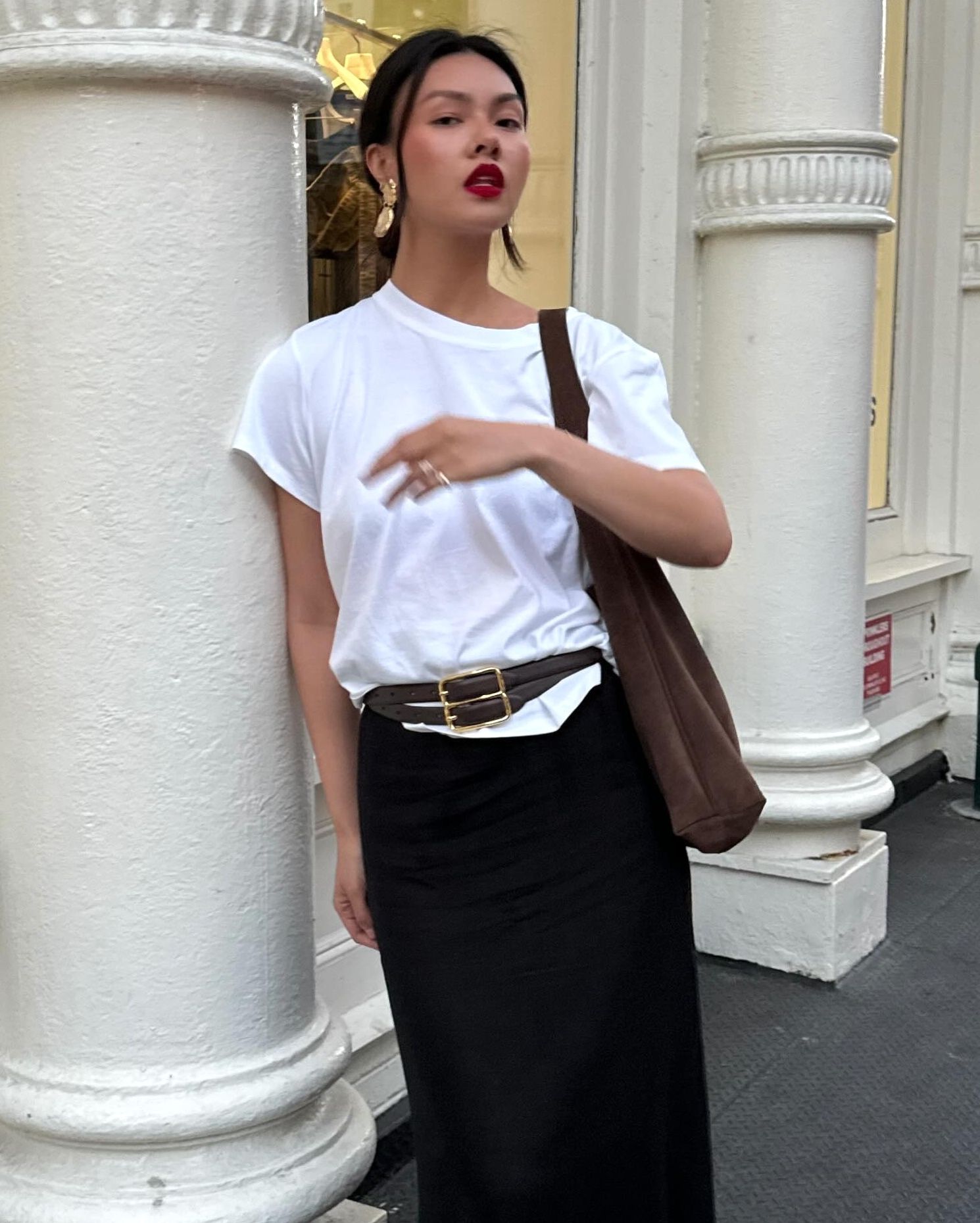 style influencer Mimi Nguyen poses on a NYC sidewalk wearing yellow-gold statement drop earrings, red lipstick, an oversize white T-shirt, brown suede tote bag, double strap buckle belt, and black skirt