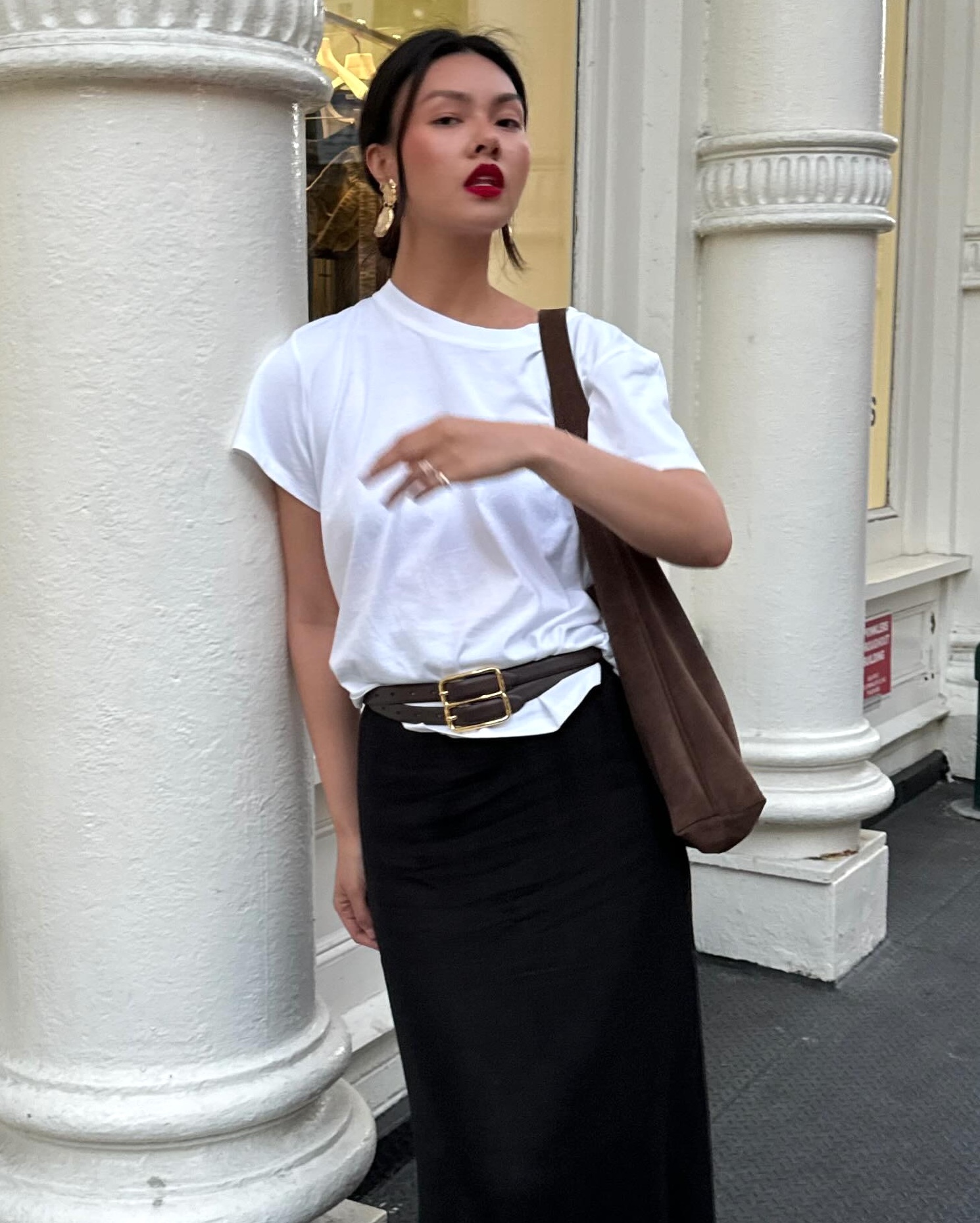 style influencer Mimi Nguyen poses on a NYC sidewalk wearing yellow-gold statement drop earrings, red lipstick, an oversize white T-shirt, brown suede tote bag, double strap buckle belt, and black skirt