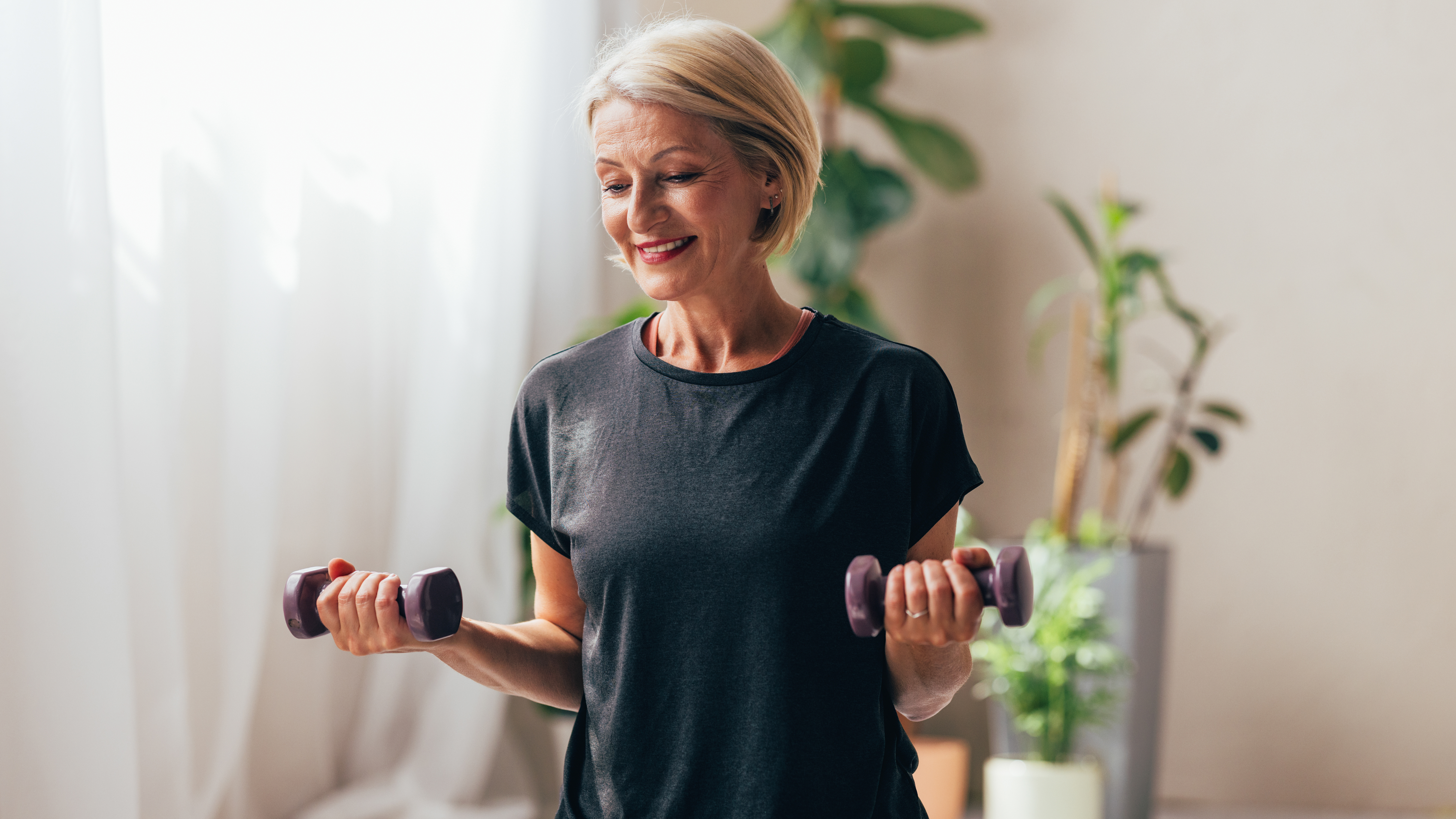 A woman stands holding lightweight dumbbells in each hand, with her elbows bent and held close to her sides. Behind her we see a curtain and leafy plants.