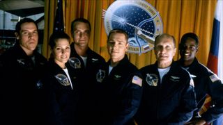 From left to right, the cast of Deep Impact (1998): Jon Favreau, Mary McCormack, Aleksandr Baluev, Ron Eldard, Robert Duvall, and Blair Underwood. All are wearing a dark blue space jumpsuit and standing in front of an orange curtain and a space emblem.