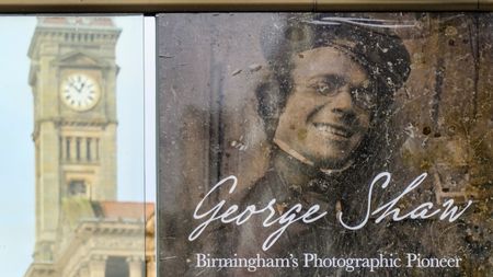 A slightly worn, sepia-toned image of a smiling, bespectacled man with the text "George Shaw: Birmingham's Photographic Pioneer," partially obscuring a blurred, tall clock tower in the background.
