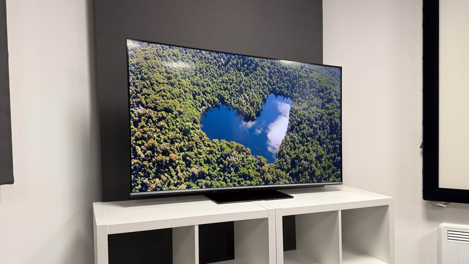 Mitchell &amp;amp; Brown QLED1811 QLED TV on white shelving unit, on screen is aerial view of heart-shaped lake