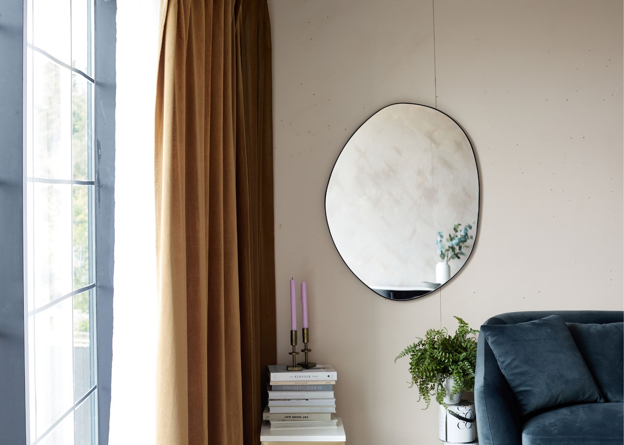 corner of a living room with neutral wall and wall mirror with sculptural shape, teal sofa in the corner