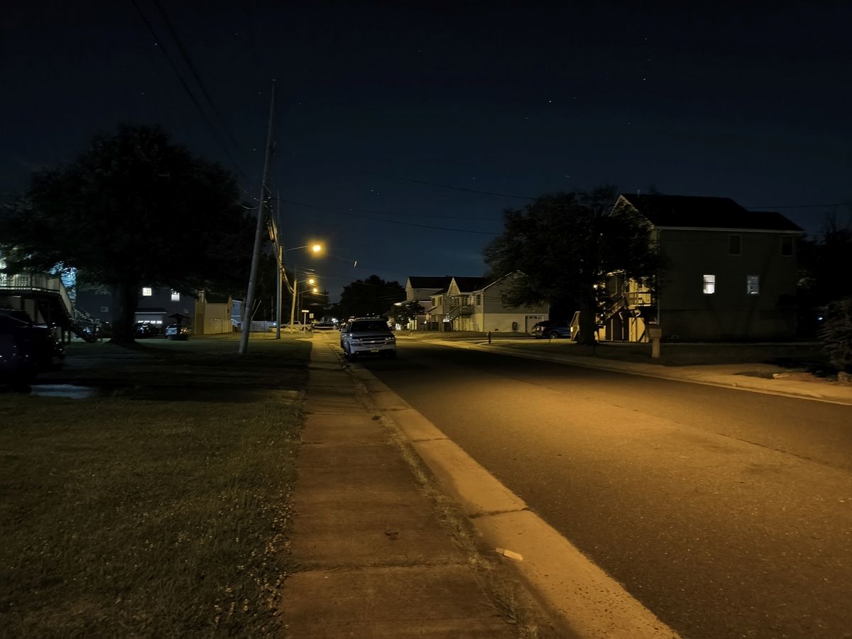 Dark suburban street late at night with no cars or people and large trees as shadows with some of the street partially illuminated with artificial light