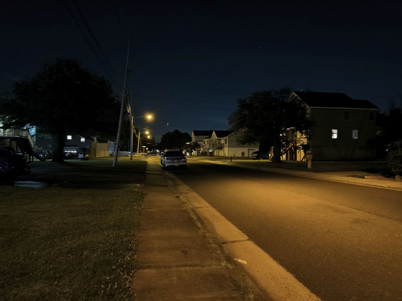 Dark suburban street late at night with no cars or people and large trees as shadows with some of the street partially illuminated with artificial light