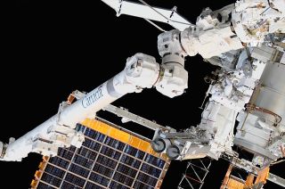 a large robotic arm in front the solar arrays of the space station. two astronauts are visible nearby in spacesuits