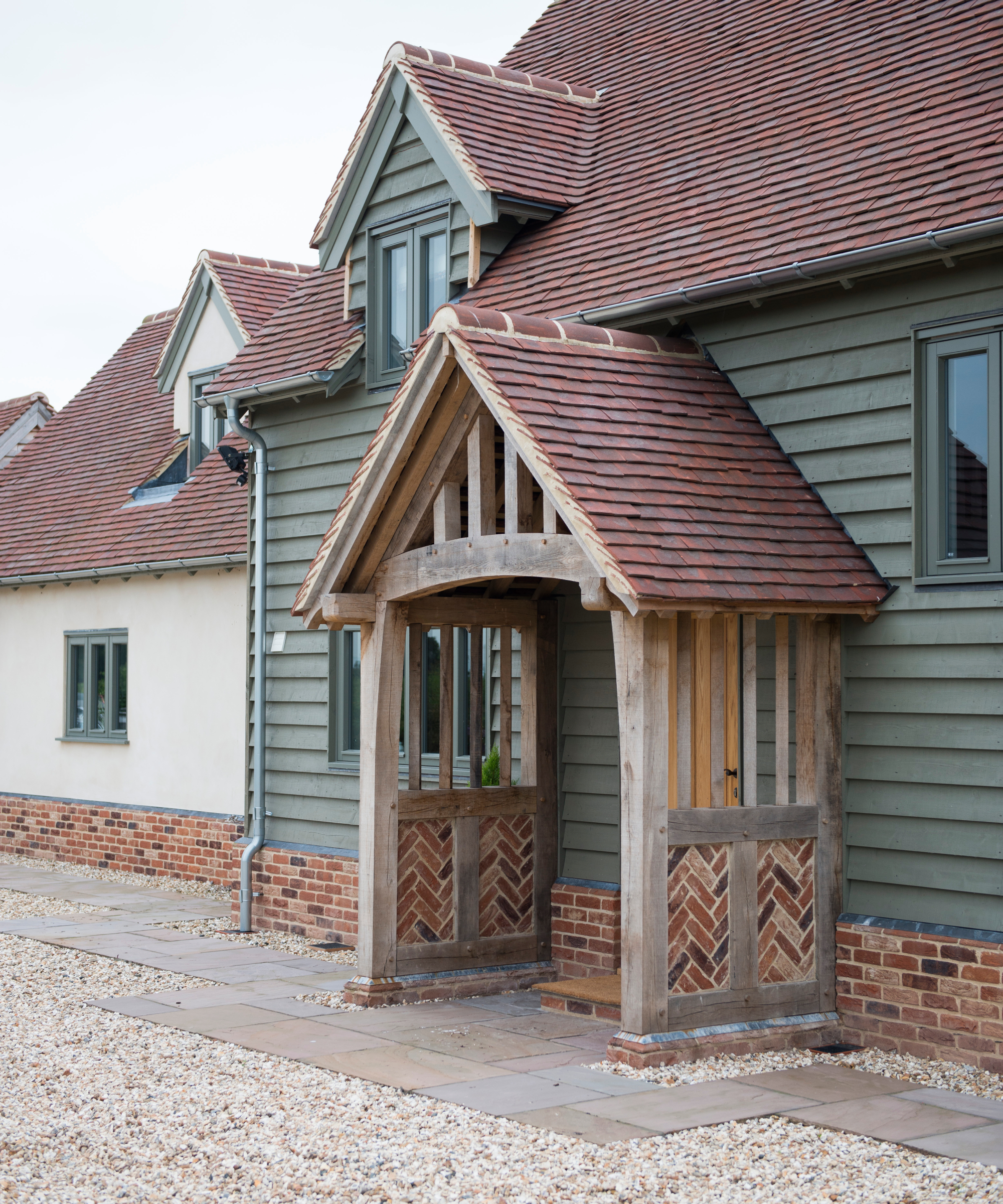 house finished with cream render, green timber cladding, oak framed porch with tiled pitched roof and lower side sections inlaid with brick in herringbone pattern