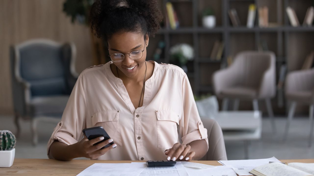 A teenage girl looks at her phone while also using a calculator at a table.