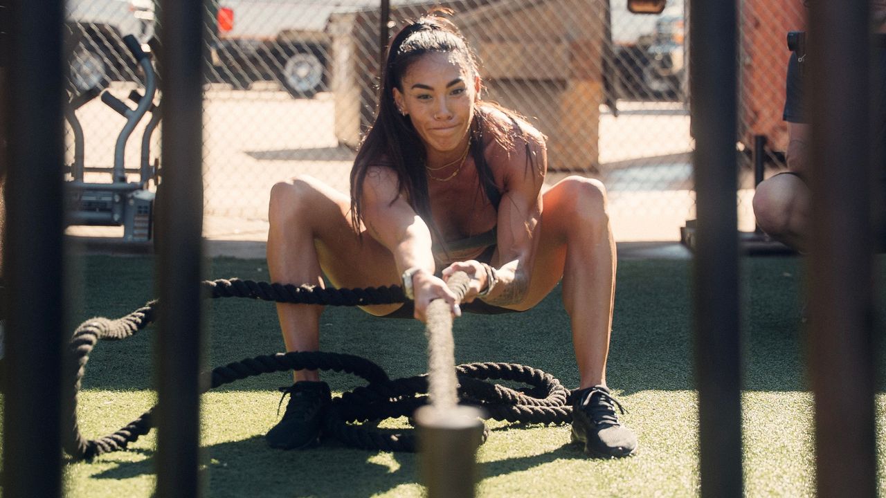 Hannah Eden, CrossFit athlete, pulls a heavy weight on a rope towards herself, while holding a squat position.