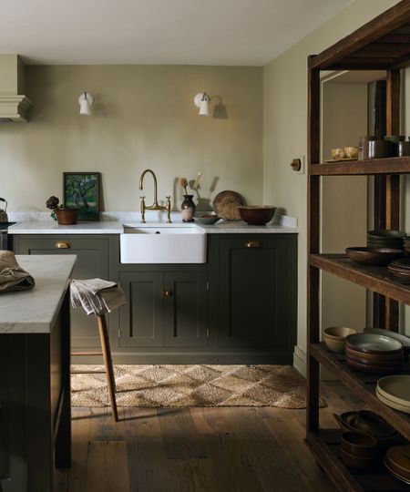 dark green kitchen cabinets and pale green walls, wooden shelving unit with tableware, wooden floorboards, marble countertop, butler sink, brass faucet, white wall lights, island, stools, rug