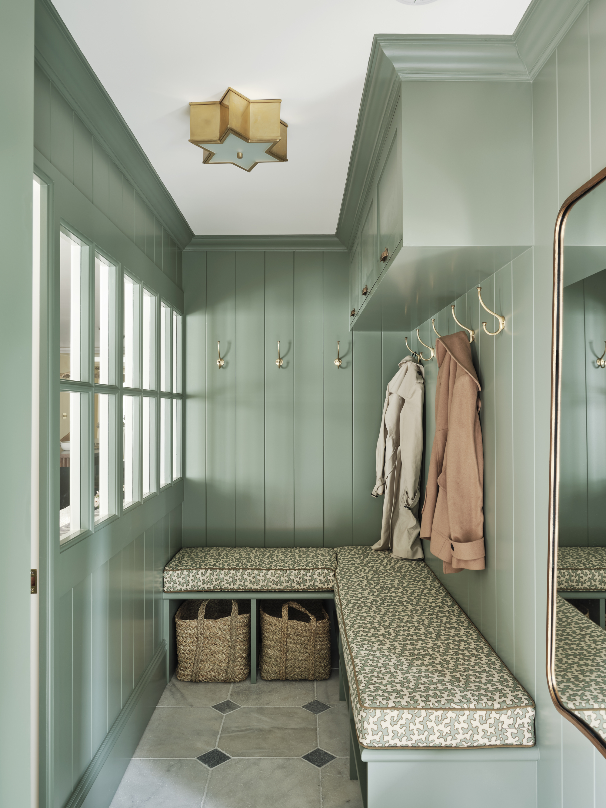 a sage green mud room with wall panelling, glass partition door, built-in banquette seat with upholstered patterned seat cushion, hooks for coats, white ceiling with star-shaped pendant light and classic tiled floors
