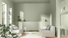 Image of a modern living room drenched in a light sage green paint color. There is light coming through the windows and shining on the wall.
