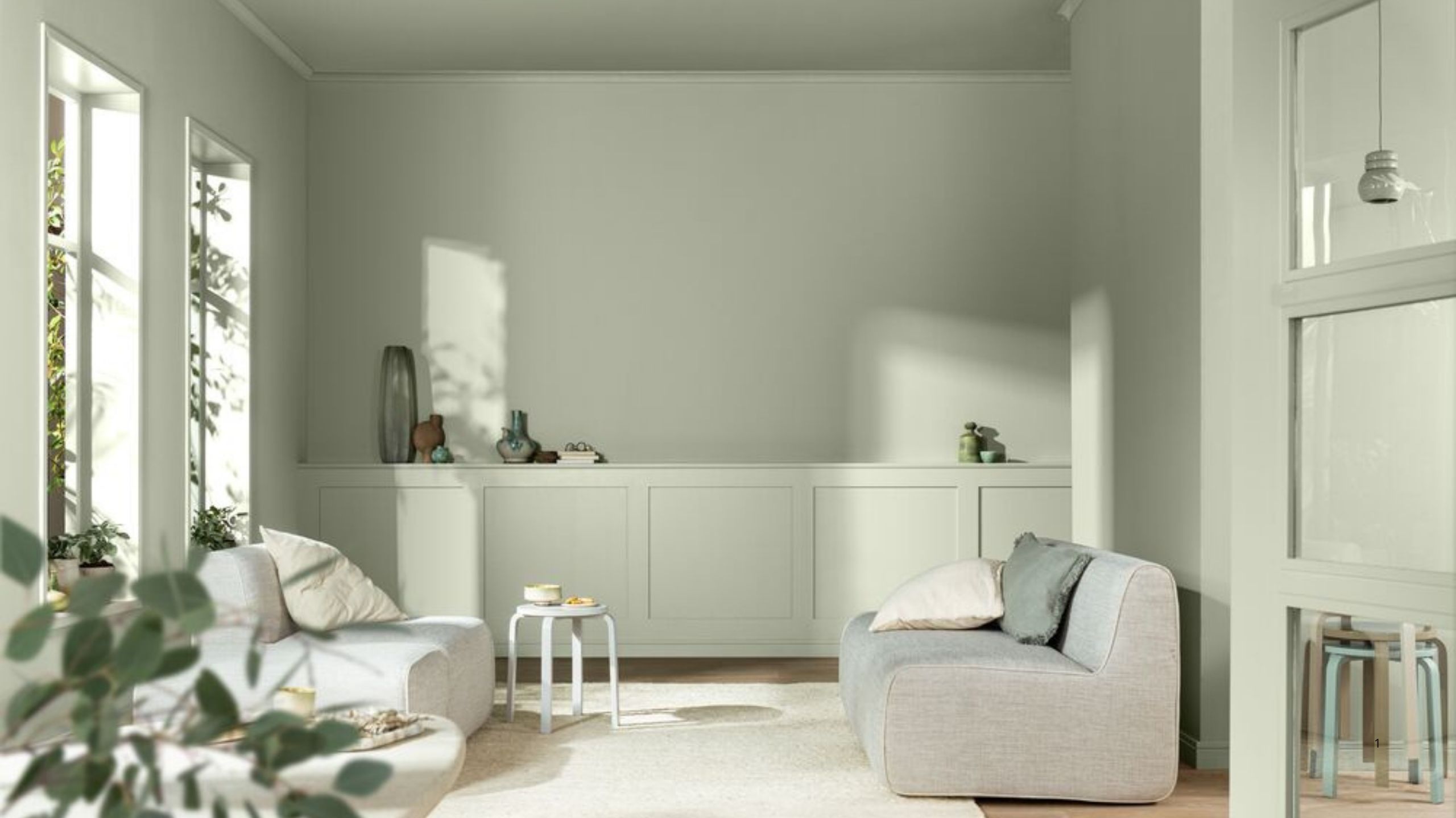 Image of a modern living room drenched in a light sage green paint color. There is light coming through the windows and shining on the wall.