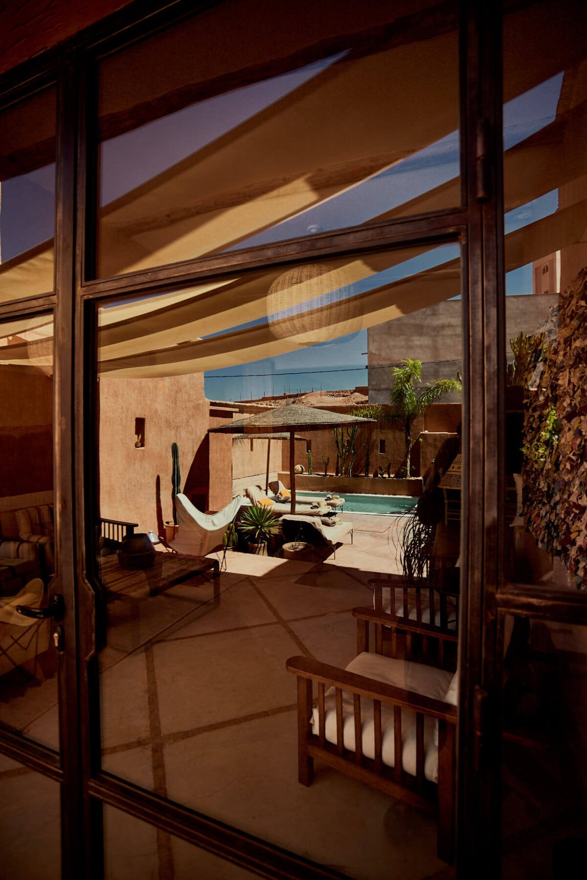 A pool patio carved from terracotta-tinted earth, decorated with wooden parasols and plants, reflects in a glass and wooden door.