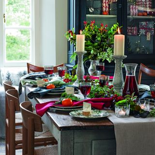 wooden dining table with wicker chairs, decorated in a country Christmas theme with green foliage with red berries, matching candles and vase, with red and blue napkins