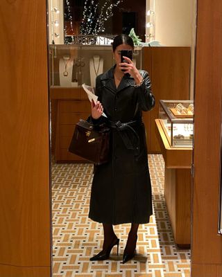 Sasha wears a leather trench coat, black sheer tights, black pumps, and a burgundy bag.