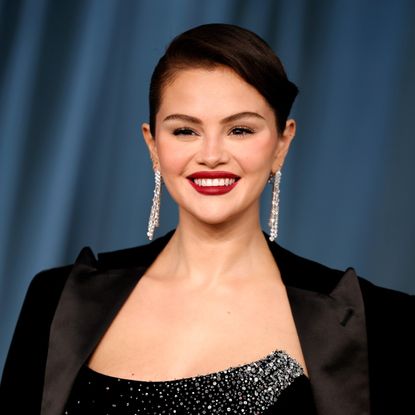 a latina woman with dark hair wearing a black blazer and posing on a red carpet in front of a blue curtain