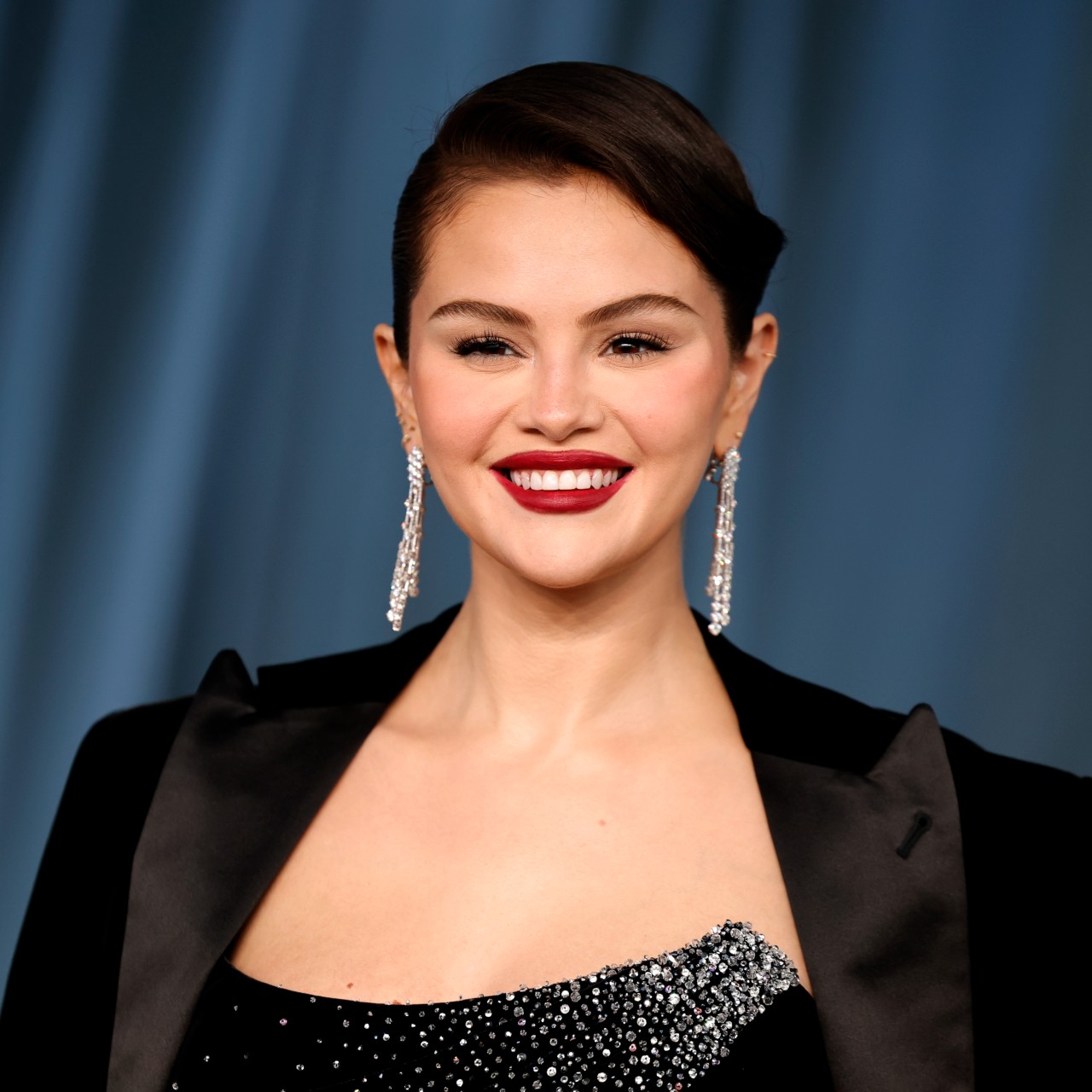 a latina woman with dark hair wearing a black blazer and posing on a red carpet in front of a blue curtain