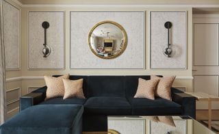 A room in the Mandarin Oriental Hyde Park. Walls with trimming are painted in light gray and beige, with a mirror and light fixtures in black. A dark blue velvet couch with a mirror-like coffee tale takes up most of the space.