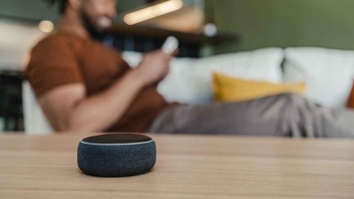 Young African American man sitting on the couch and looking at his smart phone. Smart speaker is placed on a table in the living room.